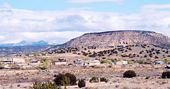 [In the far distance on the left are some snow-capped mountains. In the foreground are homes built along flat ground with little vegetation around. In the mid-distance is a tall mesa with some green shrubbery growing at lower levels giving dots of color to the light brown ground and rock. One edge of the mesa is in the middle of the image is a sloping section from the ground to the top.]